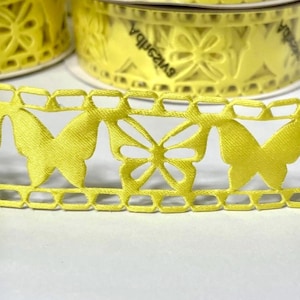 May include: Close-up of a bright yellow ribbon with a butterfly cut-out design. The ribbon is on a roll, with a second roll visible in the background. The ribbon has a delicate, openwork pattern, perfect for crafts and embellishments.