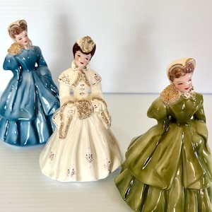 May include: Three ceramic figurines of women in vintage dresses. The dresses are in shades of blue, white, and green. Each figurine has a hat and a gold-colored collar. The figurines are posed in a classic style.