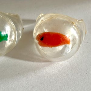 May include: Two clear, plastic rings with small, colorful fish inside. One ring contains a green fish, and the other has a bright orange fish with a black eye. The rings are set against a light background.