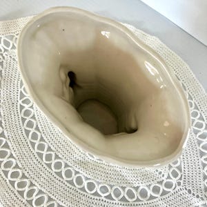 May include: Cream-colored ceramic vase with an oval opening. The vase has a textured surface and a slightly irregular rim. The interior is a darker cream color. The vase is sitting on a white crocheted doily.