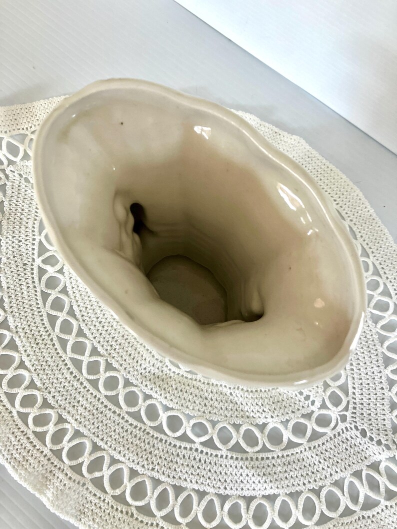 May include: Cream-colored ceramic vase with an oval opening. The vase has a textured surface and a slightly irregular rim. The interior is a darker cream color. The vase is sitting on a white crocheted doily.