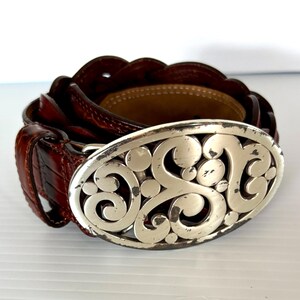 May include: A brown leather belt with a silver oval buckle. The buckle features an ornate, scrolling design. The belt is made of textured leather and has a classic, timeless style.