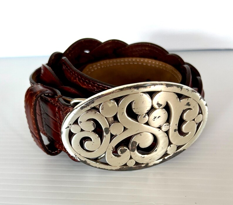 May include: A brown leather belt with a silver oval buckle. The buckle features an ornate, scrolling design. The belt is made of textured leather and has a classic, timeless style.