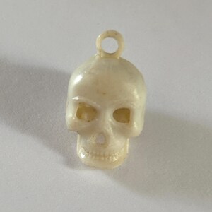 May include: A small, off-white skull charm featuring a molded loop at the top for attachment. The skull has hollow eye sockets, a nasal cavity, and detailed teeth. The surface has a smooth, slightly glossy finish with subtle variations in color.
