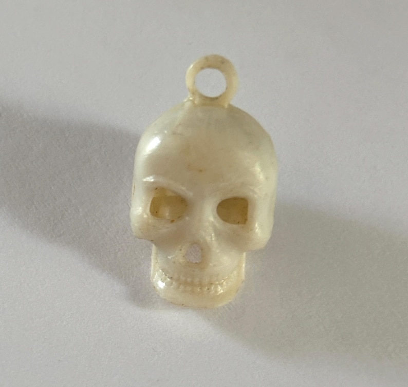 May include: A small, off-white skull charm featuring a molded loop at the top for attachment. The skull has hollow eye sockets, a nasal cavity, and detailed teeth. The surface has a smooth, slightly glossy finish with subtle variations in color.
