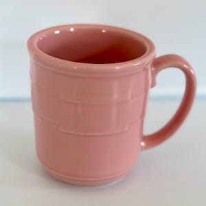 May include: A pink ceramic mug with a basket weave pattern. The mug has a rounded handle.