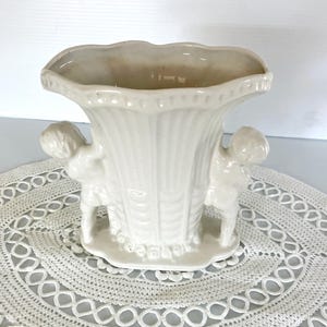May include: Off-white ceramic vase with a scalloped rim and two cherub figures on the sides. The vase has a textured, ribbed design and sits on a small, round base. The vase is displayed on a white, lace doily.