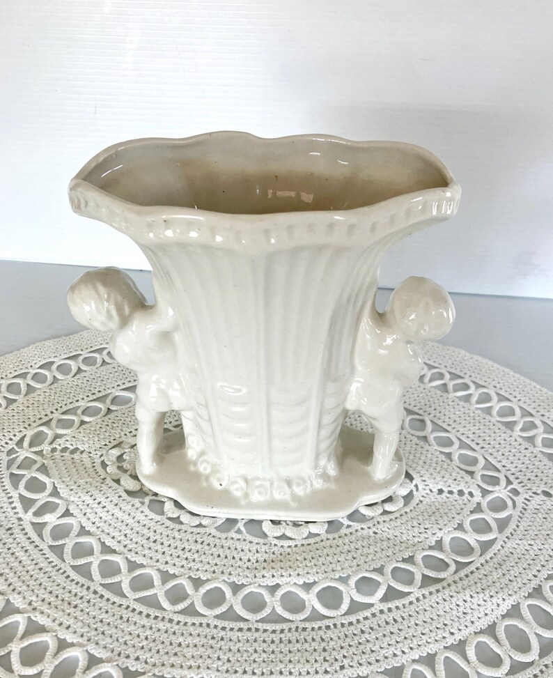 May include: Off-white ceramic vase with a scalloped rim and two cherub figures on the sides. The vase has a textured, ribbed design and sits on a small, round base. The vase is displayed on a white, lace doily.