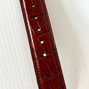 May include: Close-up of a brown leather belt with a crocodile skin texture. The belt has several evenly spaced holes and a stitched border. The leather has a glossy finish and a rich, warm color.