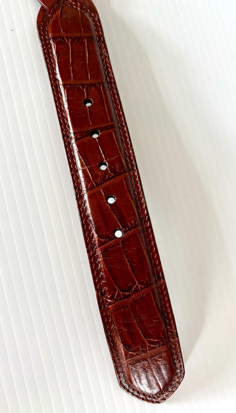 May include: Close-up of a brown leather belt with a crocodile skin texture. The belt has several evenly spaced holes and a stitched border. The leather has a glossy finish and a rich, warm color.