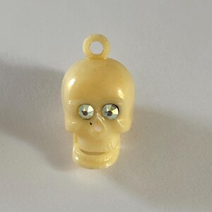 May include: A cream-colored skull charm featuring a small circular loop at the top for attachment. The skull has two faceted, clear, iridescent rhinestones set into the eye sockets and a small, smooth, light-colored nose detail.