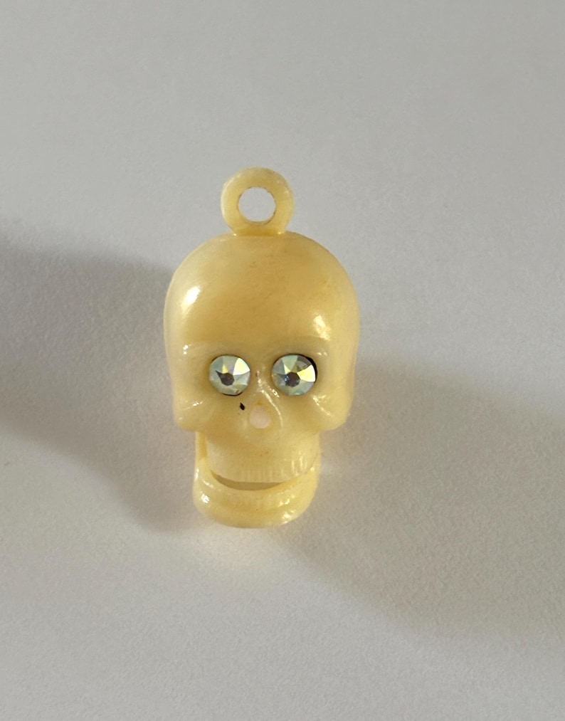 May include: A cream-colored skull charm featuring a small circular loop at the top for attachment. The skull has two faceted, clear, iridescent rhinestones set into the eye sockets and a small, smooth, light-colored nose detail.