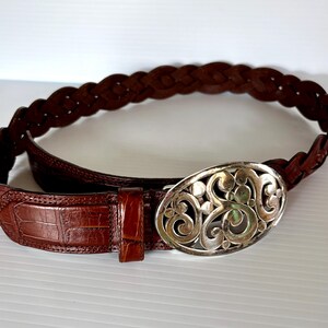 May include: A brown leather belt with a braided design and a silver oval buckle. The belt features a crocodile-textured leather section near the buckle. The buckle has an ornate, swirling pattern.