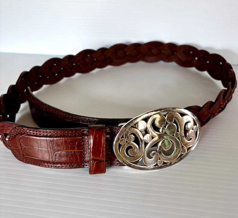 May include: A brown leather belt with a braided design and a silver oval buckle. The belt features a crocodile-textured leather section near the buckle. The buckle has an ornate, swirling pattern.