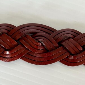May include: Close-up of a braided leather belt in a rich, reddish-brown hue. The belt's woven design is tightly constructed, showcasing the texture and detail of the leather. The belt is a classic accessory.