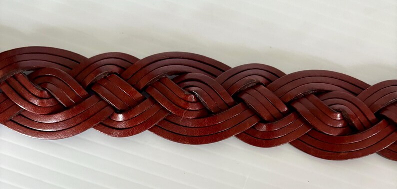 May include: Close-up of a braided leather belt in a rich, reddish-brown hue. The belt's woven design is tightly constructed, showcasing the texture and detail of the leather. The belt is a classic accessory.