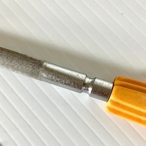 May include: A silver metal file with a yellow plastic handle. The file has a textured surface and a smooth, rounded end.