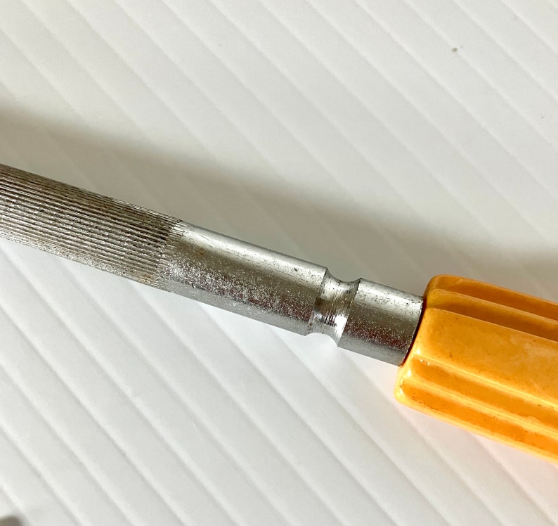 May include: A silver metal file with a yellow plastic handle. The file has a textured surface and a smooth, rounded end.