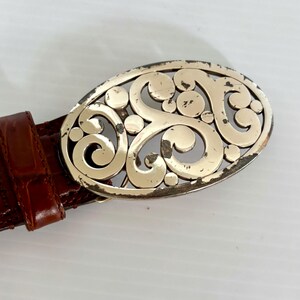 May include: A brown leather belt with a decorative silver-colored oval buckle. The buckle features an openwork design of swirling patterns and circular accents. The belt has a textured, crocodile-like pattern.