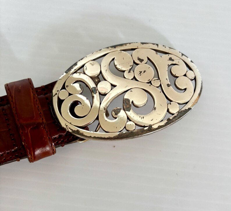 May include: A brown leather belt with a decorative silver-colored oval buckle. The buckle features an openwork design of swirling patterns and circular accents. The belt has a textured, crocodile-like pattern.