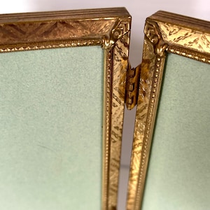 May include: A gold-colored, hinged picture frame with a light green background. The frame has ornate detailing and a decorative pattern along the edges. The hinge is visible, connecting the two sections of the frame.