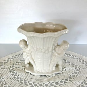 May include: Cream-colored ceramic vase with a scalloped rim and two cherub figures. The vase has a fluted body and a decorative band of roses. The vase is approximately 7 inches tall.