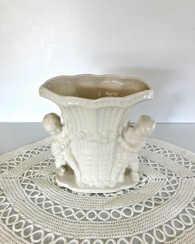 May include: Cream-colored ceramic vase with a scalloped rim and two cherub figures. The vase has a fluted body and a decorative band of roses. The vase is approximately 7 inches tall.