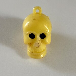 May include: A pale yellow plastic skull charm featuring a small loop at the top for hanging. The skull has two hollow, dark-colored eye sockets and a small nasal cavity opening. The surface of the charm shows minor signs of wear and texture.