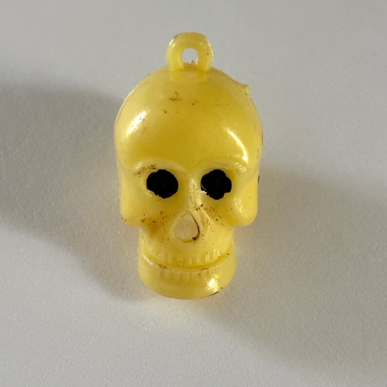 May include: A pale yellow plastic skull charm featuring a small loop at the top for hanging. The skull has two hollow, dark-colored eye sockets and a small nasal cavity opening. The surface of the charm shows minor signs of wear and texture.