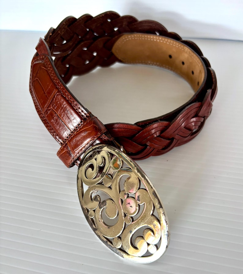 May include: A brown leather belt with a braided design and a decorative silver-colored buckle. The belt features a crocodile-textured leather section near the buckle. The buckle has an ornate, swirling pattern.