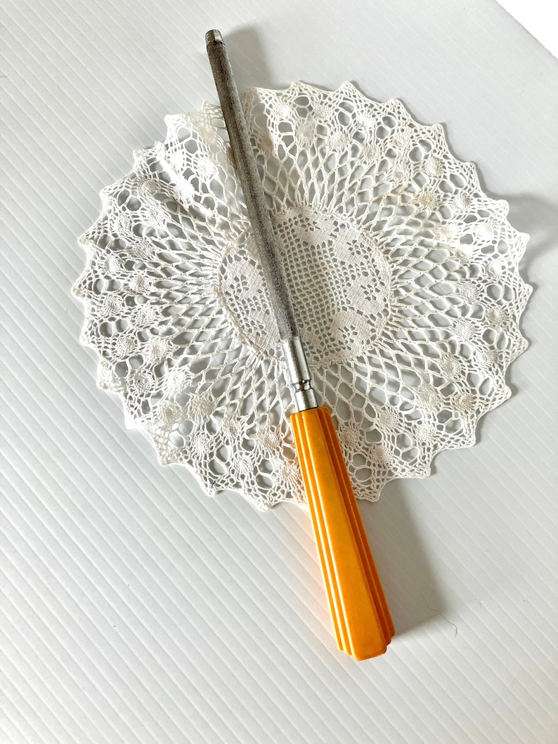 May include: A vintage knife sharpener with a yellow plastic handle and a metal blade. The sharpener is sitting on a white crocheted doily.