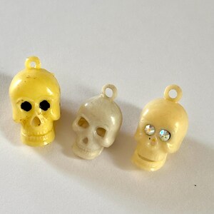 May include: Three small skull-shaped charms with loops at the top. The left charm is bright yellow with black eye sockets. The middle charm is off-white. The right charm is cream-colored and features two small, sparkling, iridescent rhinestones.