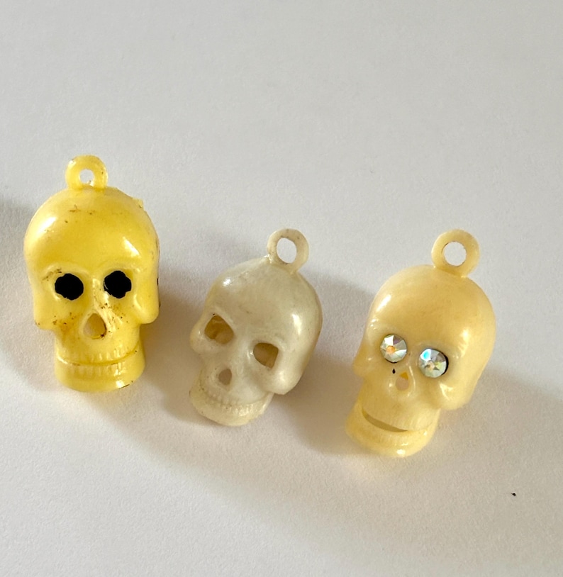 May include: Three small skull-shaped charms with loops at the top. The left charm is bright yellow with black eye sockets. The middle charm is off-white. The right charm is cream-colored and features two small, sparkling, iridescent rhinestones.