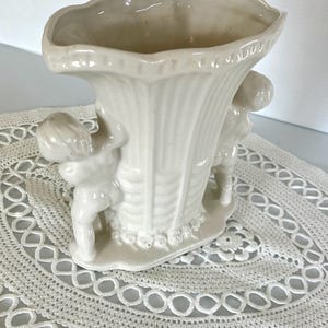 May include: Off-white ceramic vase with a fluted design and two cherubic figures. The vase has a wide, scalloped rim and sits on a small base with floral detailing. A decorative piece for home decor.