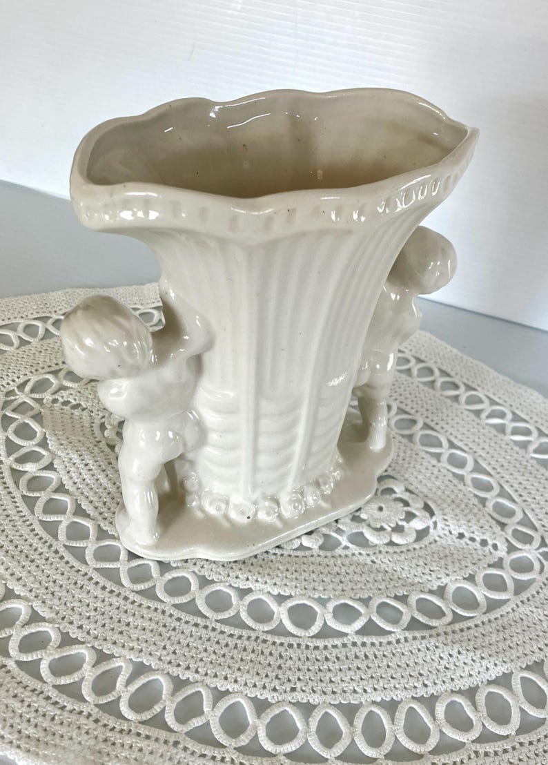 May include: Off-white ceramic vase with a fluted design and two cherubic figures. The vase has a wide, scalloped rim and sits on a small base with floral detailing. A decorative piece for home decor.