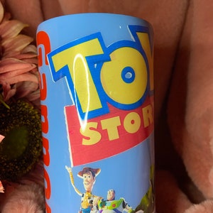 May include: A blue and white tumbler with a Toy Story design. The tumbler features Woody, Buzz Lightyear, and Rex. The text "Toy Story" is printed in yellow and red letters.
