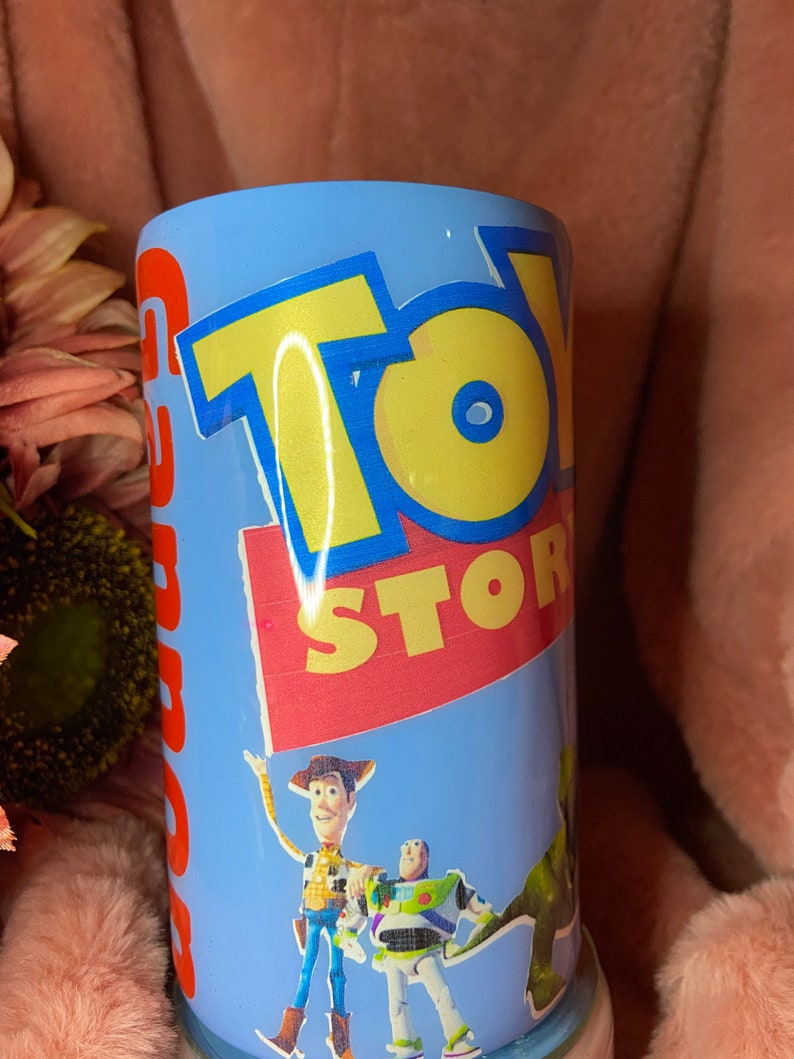 May include: A blue and white tumbler with a Toy Story design. The tumbler features Woody, Buzz Lightyear, and Rex. The text "Toy Story" is printed in yellow and red letters.
