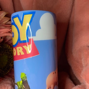 May include: A blue and white tumbler with a Toy Story theme. The tumbler features the words "Toy Story" in yellow and red letters, a white cloud, a green dinosaur, and a pink pig.