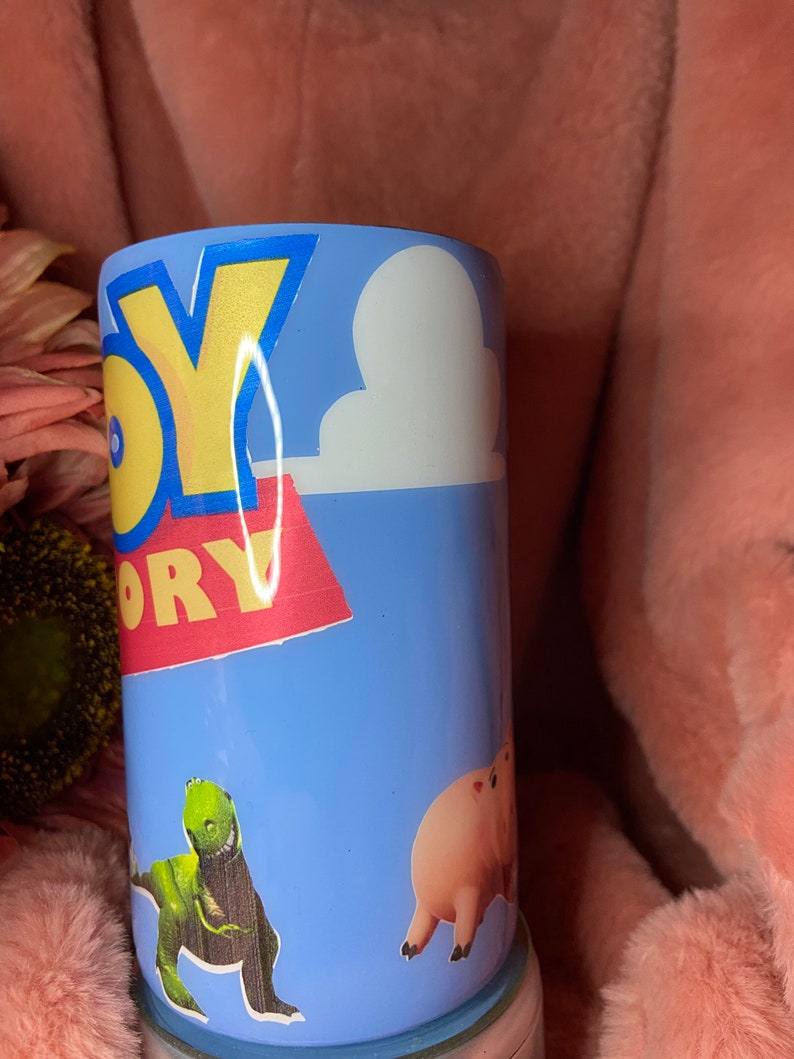 May include: A blue and white tumbler with a Toy Story theme. The tumbler features the words "Toy Story" in yellow and red letters, a white cloud, a green dinosaur, and a pink pig.