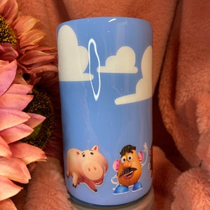 May include: A light blue tumbler with a white cloud design and characters from the Toy Story film. The characters include a pig, a potato head, and a cowboy.