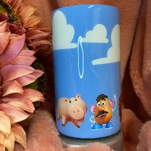 May include: A blue tumbler with a white cloud design and characters from the Toy Story film. The characters include a pig, a potato head, and a cowboy.