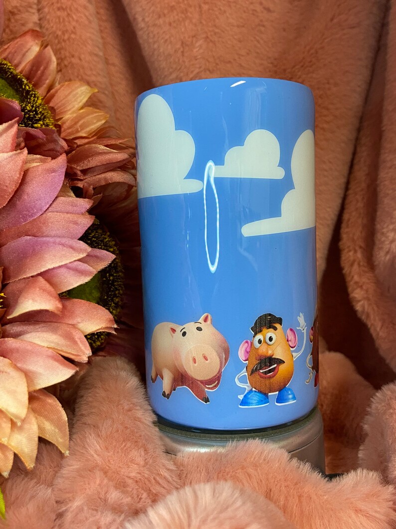 May include: A blue tumbler with a white cloud design and characters from the Toy Story film. The characters include a pig, a potato head, and a cowboy.