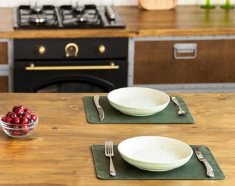 May include: A kitchen scene with a wooden table set for a meal. Four white bowls sit on green placemats, each with a fork and knife. A small glass bowl of cherries is in the center. The kitchen has wooden cabinets and a black oven.