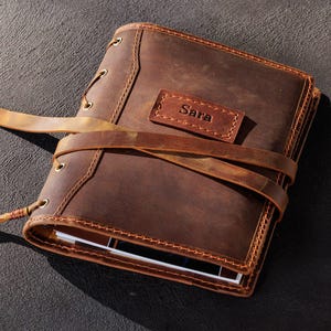 May include: A brown leather journal with the name "Sara" embossed on a small rectangular patch. The journal is secured with a leather strap and sits next to a white pencil with a green eraser.