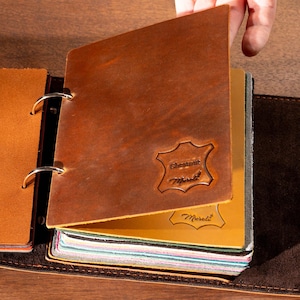 May include: Open leather journal with a brown cover and dark brown suede-like backing. The journal is open, revealing colorful pages. The cover has a debossed emblem. The journal is on a wooden surface.