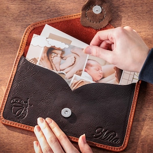 May include: A dark brown leather photo album with a light brown border, holding several photographs. The album has a magnetic closure and a decorative design. The name "Mila" is embossed on the album.