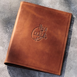 May include: A brown leather menu cover with the embossed text "Allison Cocktails" in a circular design. The cover is rectangular with rounded corners and a stitched border. The background is a mottled gray surface.