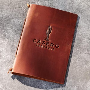 Op de afbeelding: Een bruine leren menucover met de in reliëf aangebrachte tekst "CASERO TAQUERIA" en een cactusillustratie. De cover is gesloten met een leren koord. De achtergrond is een gevlekt grijs oppervlak.
