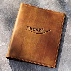 May include: A brown leather menu cover with the text "El Camino Real" and a chili pepper design embossed on the front. The cover has a textured appearance and a stitched border. The background is a mottled gray surface.