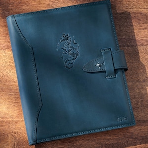 Op de afbeelding: Een donkerblauw leren notitieboek met een drakenembleem in reliëf op de voorkant. Het notitieboek heeft een gespsluiting en staat op een houten achtergrond. Het notitieboek is een notitieboek.
